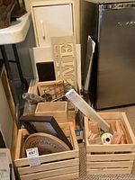 Photo of two wooden crates containing various scrap wood pieces, wooden wall signs including one with the word HOME, and other crafting supplies near a fridge.