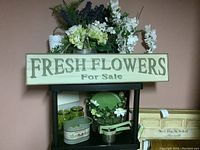 Photo showing various faux flowers arranged in different vases including a ceramic container and a metal bucket, decorative green glass jars, a green ceramic plate, and a wooden wall sign that reads 'Fresh Flowers For Sale'. Also a handheld ricer and a green metal bucket labeled 'Farmhouse' are visible.