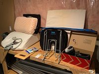 Photo of lot including beige foam wedge pillow, peach memory foam pillow, neck brace, slippers in box, metal cane stand, travel wipes, cane, and carrying bag on wooden surface with plywood and pink insulation background.