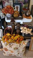 Overall view of the lot showing faux flower arrangements, votive owl figures, metal bowls, wooden candlesticks, and baskets containing faux vegetables.