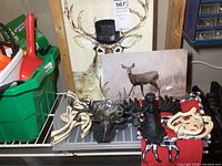 Shows black cast iron deer and moose head wall hooks, cream-colored cast iron door knockers with rust, and two deer-themed art prints featuring a stag and a doe on canvas, plus a black cast iron deer or moose sculpture figure all arranged on a shelf.