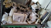 Wide shot of assorted home decor items on metal shelf including ceramic pitchers, faux florals, wrapped electric candles, canvas prints, platters, cast iron candleholder, and faux antlers.