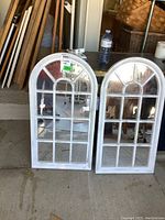 Two matching distressed white wood framed window style mirrors with arched tops and segmented panes, leaning against a wall in garage space.