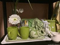 Photo showing green ceramic mugs, faux flowers, towel, small container, and part of platters on shelf.