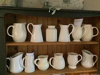 Full view of two wooden shelves with 12 white ceramic pitchers of various shapes and sizes arranged in two rows.