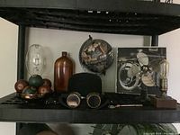 Overview of the lot displayed on black shelf including light bulb, glass jug, pool balls, boiler hat with goggles, fan, globe, pocket watch, and framed motorcycle photo.