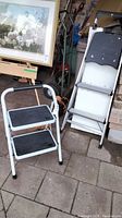 Two-step stool and folding step ladder side by side on patio against garage wall
