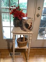 Front view showing the distressed white wood side table with a woven basket on the shelf below, rooster tin, and faux flower planter on top.