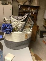 Full view of basket holding two throw pillows, satin-like fabric, framed mirror plaque, and faux flower