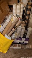Assorted wallpaper and craft paper rolls stored in bags and boxes, showing variety of patterns including checkered beige, black, stripes and neutrals.