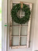 Full view of the vintage wooden window frame with peeling white paint and mounted green leaf wreath, hung on wall.