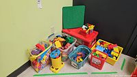 Full view of seven containers holding various blocks and toys including a yellow Playskool bucket, clear bins, and a red Duplo firetruck themed box with green base plates.