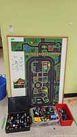 Wooden floor track with road and town layout, showing some wear and repair needed. Several bins of toy cars placed in front.