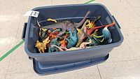 Blue plastic bin filled with assorted colored plastic dinosaur figures on a tiled floor.