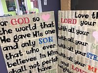 Two Biblical-themed wall hangings side by side, showing colorful text on a newspaper page background, placed indoors.