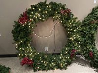 Full view of the large circular artificial Christmas wreath with green pine needles, integrated white string lights, and red berry decorations, approximately 64 inches diameter.