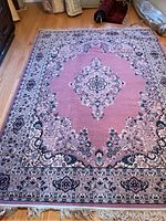 Full view of the rectangular wool pile area rug showing detailed borders and central medallion in pink tones with floral patterns and fringe edges.