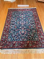 Full view of rectangular wool area rug displaying central floral pattern, deep red border, and cream-colored fringes on hardwood floor.