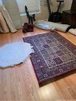 Photo showing both the red IKEA Valby Ruta rectangular rug with floral and geometric patterns and the smaller white octagonal Anglo wool rug with fringe. Also visible are rolled up rugs in the background.