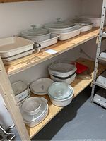 Two-level wooden shelving holding multiple white fluted ceramic dishes with glass lids on top shelf, assorted CorningWare and Pyrex pieces on bottom shelf.