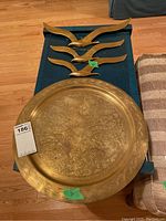 Large brass display plate showing detailed engraved patterns and decorative border with three brass bird decor pieces above it on a table.