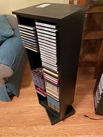Photo shows a black swivelling CD tower filled with CDs on both sides, located by a sofa and wood floor.