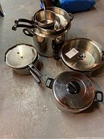 Photo showing a variety of cookware items including large and small pots, a skillet with black handles, and lids stacked on a basement floor.