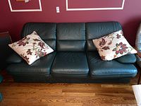 Front view of green leather three seat couch with two floral accent cushions on each end.