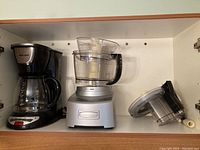 Photo showing set of kitchen appliances including black drip coffee maker and silver Cuisinart food processor with clear bowl and lid.