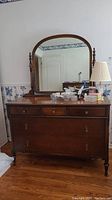 Full view of wooden dresser with attached rounded top mirror placed on hardwood floor against wallpapered wall.