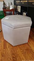 Full view of the hexagonal light grey leather storage cube/foot rest on wood floor.