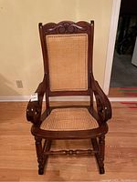 Front view of wooden cane rocking chair showing carved details on top rail, armrests and cane seat and back.
