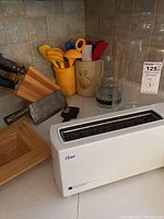 Full view of kitchen setup showing Oster Cool Touch toaster, yellow utensil holder with various spatulas, Henkels knife block, Chinese cleaver, glass jars, and ceramic holder