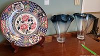 Photo showing large floral decorative plate on wooden stand and two blue gradient glass vases.