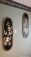 Photo showing two black lacquered oval wall plaques hanging on a pale green wall with detailed bird and floral shell inlay designs.