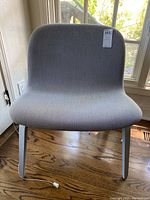 Front view of gray upholstered lounge chair showing curved seat and backrest, sitting on wood floor near window.