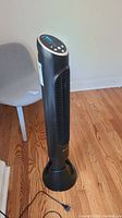 Full view of Honeywell black tower fan standing on wooden floor near a grey chair, power cord visible on the floor.