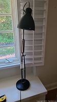 Full view of the black adjustable table lamp showing the lamp base, arm with spring tension adjustment, and lamp shade in front of a window.