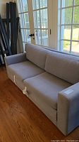 Gray fabric sofa bed tied down, with three back cushions, straight arms, and modern design on hardwood floor near large windows.