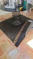 Black woven throw rug with beige fringed edges partially folded over on terracotta tile flooring. Shows use and areas needing cleaning.