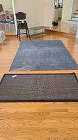 Image showing both throw rugs on wood floor: larger gray rug with stains and smaller checkered rug with black border