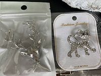 Photo showing both the silver-tone reindeer head brooch in a clear plastic bag and the cat-shaped rhinestone brooch on white card packaging.