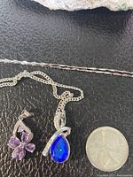 Sterling silver chains with purple flower and blue teardrop pendants, shown next to coin for scale.