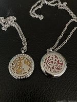 Two round silver-tone oil diffuser necklaces with decorative front covers, one with cat and butterfly design with rhinestones, the other with swirl pattern. Both have silver-colored chains.