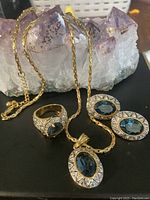 Front view of gold-tone chain necklace, pendant with large oval blue stone, matching oval blue stone earrings, and ring with blue stone missing one stone, all placed on amethyst crystal cluster.