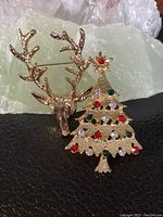 Photo showing the gold-tone Christmas tree brooch adorned with red, green and clear rhinestones alongside the matching reindeer head brooch with textured gold-tone finish and detailed antlers.