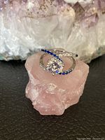 Image showing front top view of silver filled ring with large cubic zirconia center and blue and clear stone swirl accents, placed on a pink quartz crystal with an amethyst crystal background.