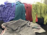 Photo showing multiple hoodies and sweaters in colors including purple, teal, red, green, and grey with zippers and drawstrings visible.