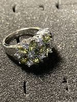 Close-up photo of silver ring with light green and clear stones arranged in a flower shape on a black textured background.