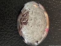 Close-up front view of oval sterling silver brooch showing detailed floral and scroll engraving on polished surface.